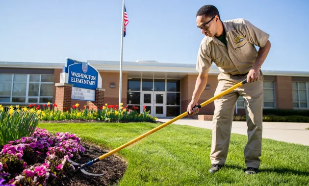 A hardworking general worker in a school uniform using gardening tools to trim grass and maintain flower beds, with a clean and bright school building in the background. Include elements like school signage, bright sunlight, and a peaceful educational environment.