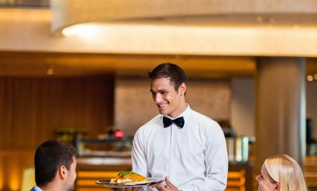 A professional hotel restaurant setting with a smiling waiter serving food to well-dressed guests. The environment should feel elegant and welcoming, with modern décor and warm lighting. Waiter should look confident, neat, and friendly