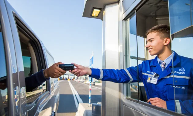 A smiling young adult tollgate cashier standing at a modern toll plaza, wearing a uniform and assisting a driver, with bright skies in the background. The image should be friendly, inviting, and professionally designed with bold colors to catch attention on mobile screens.
