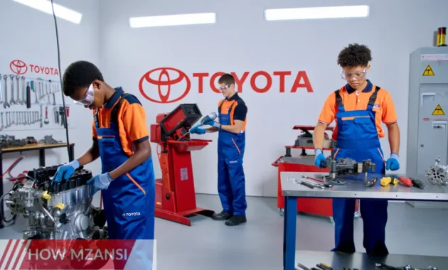 an image of young apprentices in a Toyota-branded workshop wearing overalls and safety gear, working on car engines and industrial machines. Include visible tools, electrical panels, and mechanical parts to show hands-on learning. The background should be bright and clean, with Toyota branding on the walls.